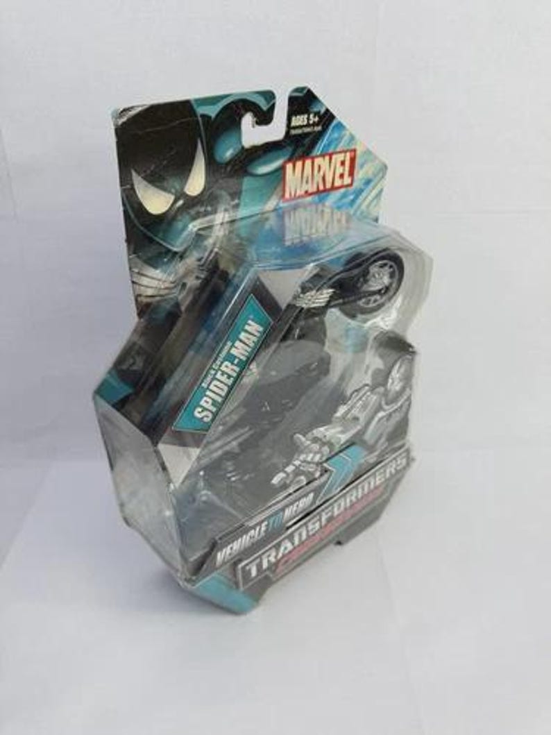 Puede incluir: Una figura de acci&oacute;n de Marvel Spider-Man empaquetada. El embalaje es azul y negro con el logotipo de Marvel y las palabras "Spider-Man" y "Vehicle to Hero Transformers". La figura es una motocicleta.