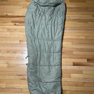 May include: An olive green sleeping bag, laid out on a wooden floor. The bag is long and tubular, with a hood at the top and horizontal stitching across its length. It appears to be made of a lightweight, possibly water-resistant material, suitable for camping or outdoor use.