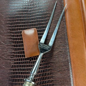 May include: A vintage carving fork with a stainless steel blade and a decorative handle. The handle has a silver-colored metal bolster and a natural-colored, textured grip. The fork is marked "BROWN & BIGELOW SOLINGEN GERMANY".