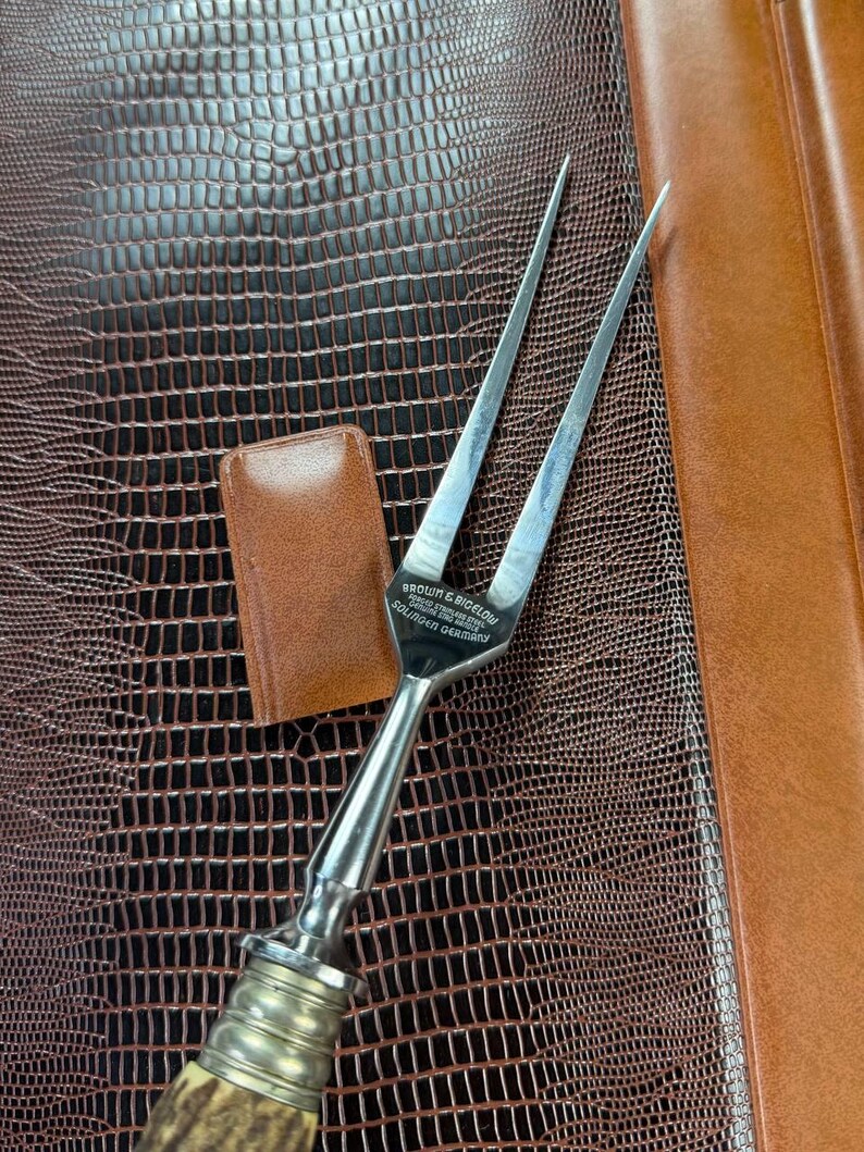 May include: A vintage carving fork with a stainless steel blade and a decorative handle. The handle has a silver-colored metal bolster and a natural-colored, textured grip. The fork is marked "BROWN & BIGELOW SOLINGEN GERMANY".