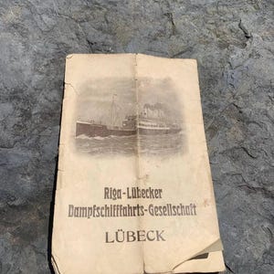 Op de afbeelding: Een vintage brochure met een sepia-getinte illustratie van een stoomschip. De omslag toont de tekst "Riga-Lübecker Dampfschiffahrts-Gesellschaft LÜBECK" in een klassiek schreeflettertype. Het papier heeft een versleten, verouderde uitstraling.