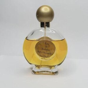 May include: A round glass perfume bottle with a gold-colored cap and a clear base. The bottle contains a golden-yellow liquid. The label reads "Eau de Toilette Bal a Versailles Jean Desprez".