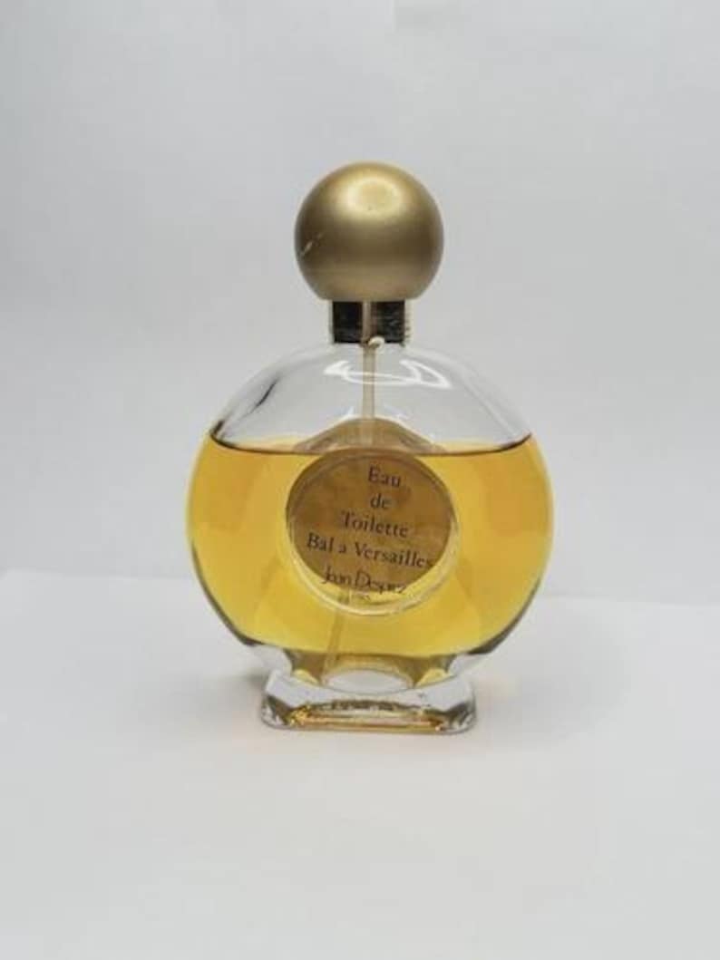 May include: A round glass perfume bottle with a gold-colored cap and a clear base. The bottle contains a golden-yellow liquid. The label reads "Eau de Toilette Bal a Versailles Jean Desprez".