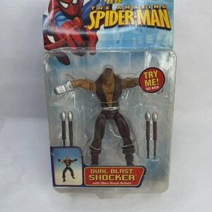 May include: The Amazing Spider-Man action figure of Dual Blast Shocker in a sealed package. The figure is brown and tan with silver accessories. The packaging features Spider-Man graphics and the Marvel logo.