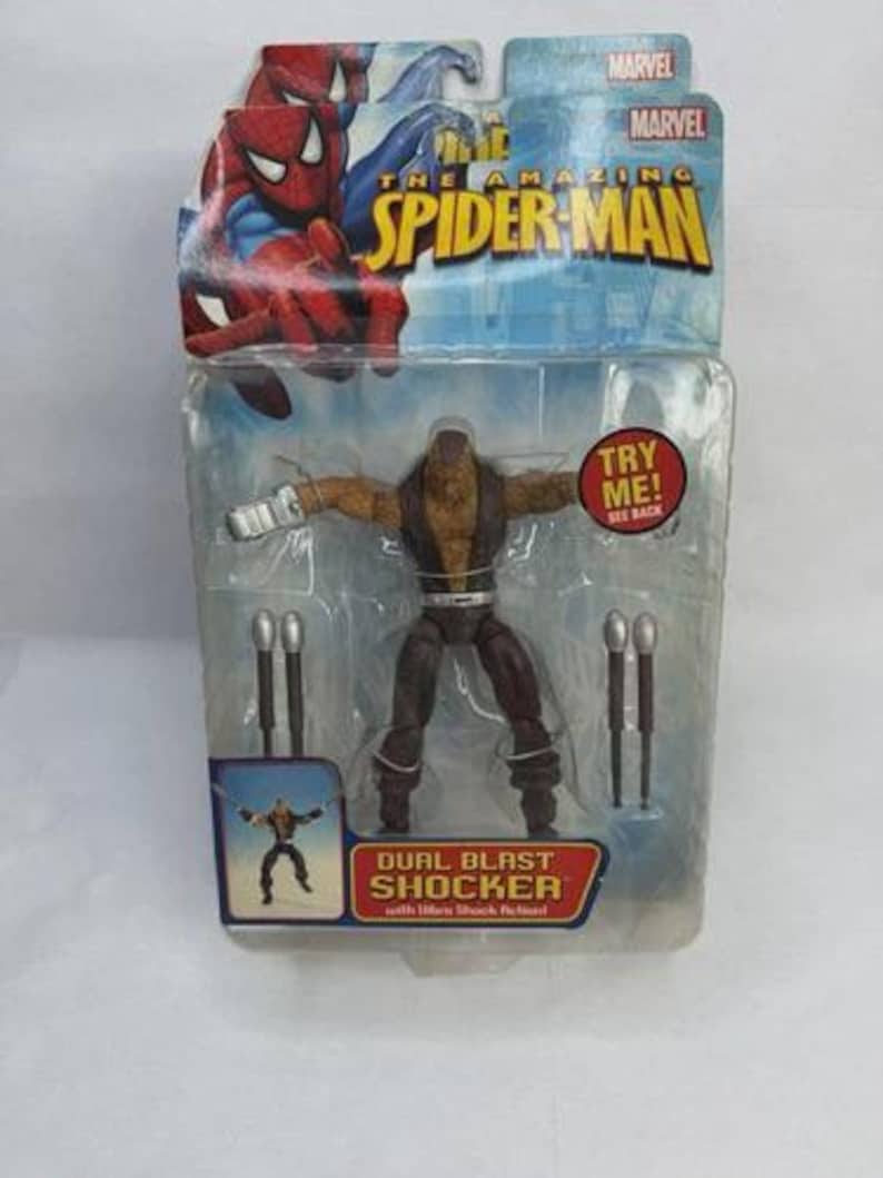 May include: The Amazing Spider-Man action figure of Dual Blast Shocker in a sealed package. The figure is brown and tan with silver accessories. The packaging features Spider-Man graphics and the Marvel logo.