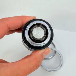 Puede incluir: Un objetivo de c&aacute;mara plateado y negro con un elemento de vidrio transparente. Un anillo plateado con el texto "Germany" y "1527" est&aacute; junto al objetivo. El objetivo se sostiene en una mano sobre un fondo blanco.