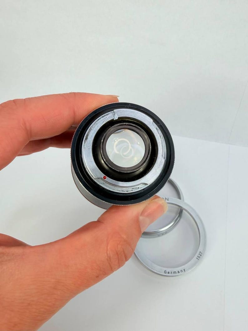 Puede incluir: Un objetivo de c&aacute;mara plateado y negro con un elemento de vidrio transparente. Un anillo plateado con el texto "Germany" y "1527" est&aacute; junto al objetivo. El objetivo se sostiene en una mano sobre un fondo blanco.