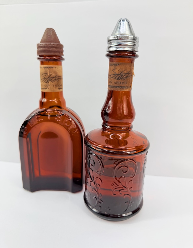 Puede incluir: Dos botellas vintage de vidrio &aacute;mbar. Una es rectangular con tap&oacute;n marr&oacute;n, la otra redonda con tapa plateada y dise&ntilde;o floral en relieve. Ambas tienen etiquetas que dicen "Stetson".