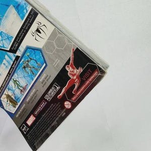 Puede incluir: Paquete parcialmente visible de una figura de acci&oacute;n de Spider-Man. El embalaje presenta un gr&aacute;fico de Spider-Man rojo y negro, un s&iacute;mbolo de ara&ntilde;a y texto que incluye "Marvel" y "Columbia Pictures". El paquete est&aacute; en &aacute;ngulo, mostrando el lateral y la parte superior.
