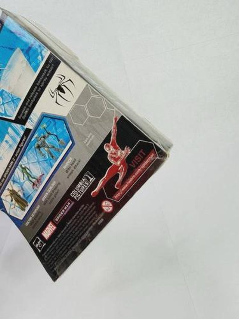 Puede incluir: Paquete parcialmente visible de una figura de acci&oacute;n de Spider-Man. El embalaje presenta un gr&aacute;fico de Spider-Man rojo y negro, un s&iacute;mbolo de ara&ntilde;a y texto que incluye "Marvel" y "Columbia Pictures". El paquete est&aacute; en &aacute;ngulo, mostrando el lateral y la parte superior.