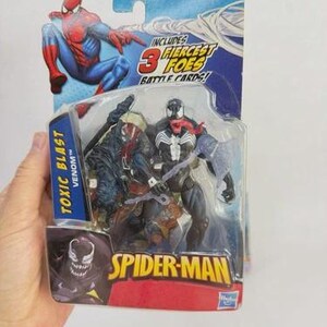 May include: A Spider-Man action figure set in a sealed package. The package features Spider-Man, Venom, and other characters. The text "TOXIC BLAST VENOM" and "SPIDER-MAN" are visible. The packaging includes the text "INCLUDES 3 FIERCEST FOES BATTLE CARDS!"