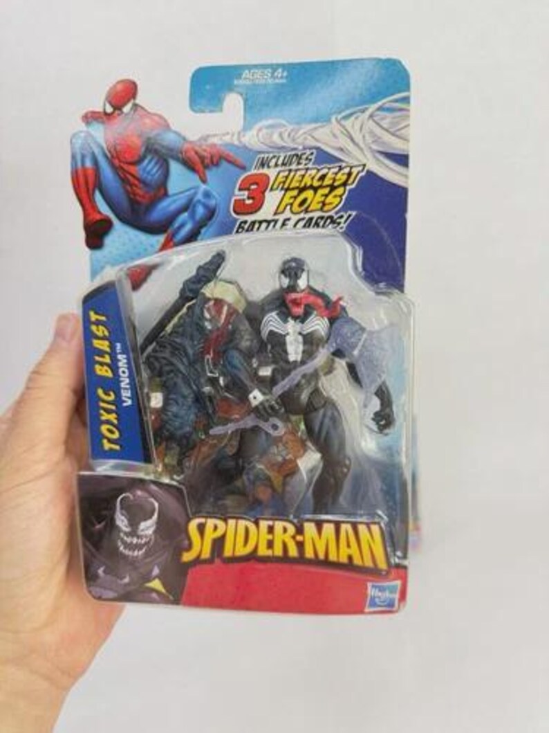 May include: A Spider-Man action figure set in a sealed package. The package features Spider-Man, Venom, and other characters. The text "TOXIC BLAST VENOM" and "SPIDER-MAN" are visible. The packaging includes the text "INCLUDES 3 FIERCEST FOES BATTLE CARDS!"