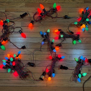 May include: A collection of multi-colored string lights featuring opaque bulbs in red, orange, green, and blue. The lights are connected by dark brown electrical cords and include black plastic sockets, arranged in several tangled clusters.