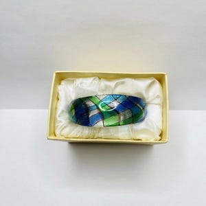 May include: A patterned bracelet in a small yellow gift box. The bracelet features a mosaic design in blue, green, and silver. The box is lined with white satin. The bracelet's design is geometric.