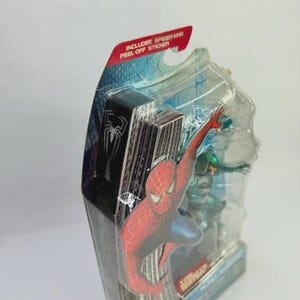 Puede incluir: Figura de acci&oacute;n de Spider-Man en un paquete de pl&aacute;stico transparente. La figura es roja y azul, con un tel&oacute;n de fondo de paisaje urbano. El paquete incluye una pegatina despegable.