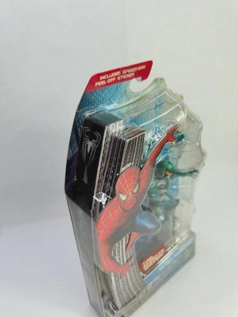 Puede incluir: Figura de acci&oacute;n de Spider-Man en un paquete de pl&aacute;stico transparente. La figura es roja y azul, con un tel&oacute;n de fondo de paisaje urbano. El paquete incluye una pegatina despegable.