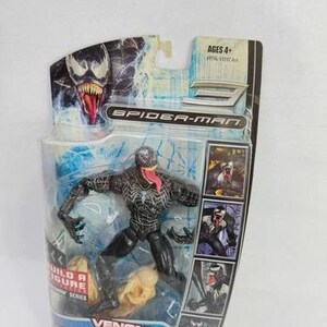 May include: A Venom action figure from the Spider-Man series, packaged in a clear plastic blister pack. The figure is black with white detailing and a red tongue. The packaging includes the text "Venom" and "Collect all eight figures."