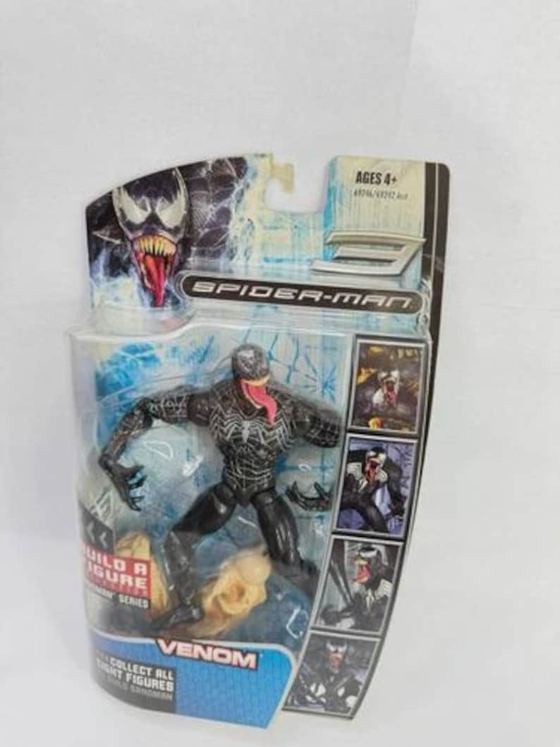 May include: A Venom action figure from the Spider-Man series, packaged in a clear plastic blister pack. The figure is black with white detailing and a red tongue. The packaging includes the text "Venom" and "Collect all eight figures."