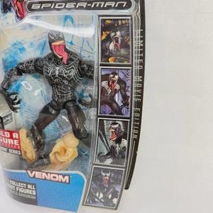 May include: A Venom action figure from the Spider-Man movie series, in its original packaging. The figure is black with white accents and a long red tongue. The packaging includes the text "VENOM" and "COLLECT ALL FIGHT FIGURES".