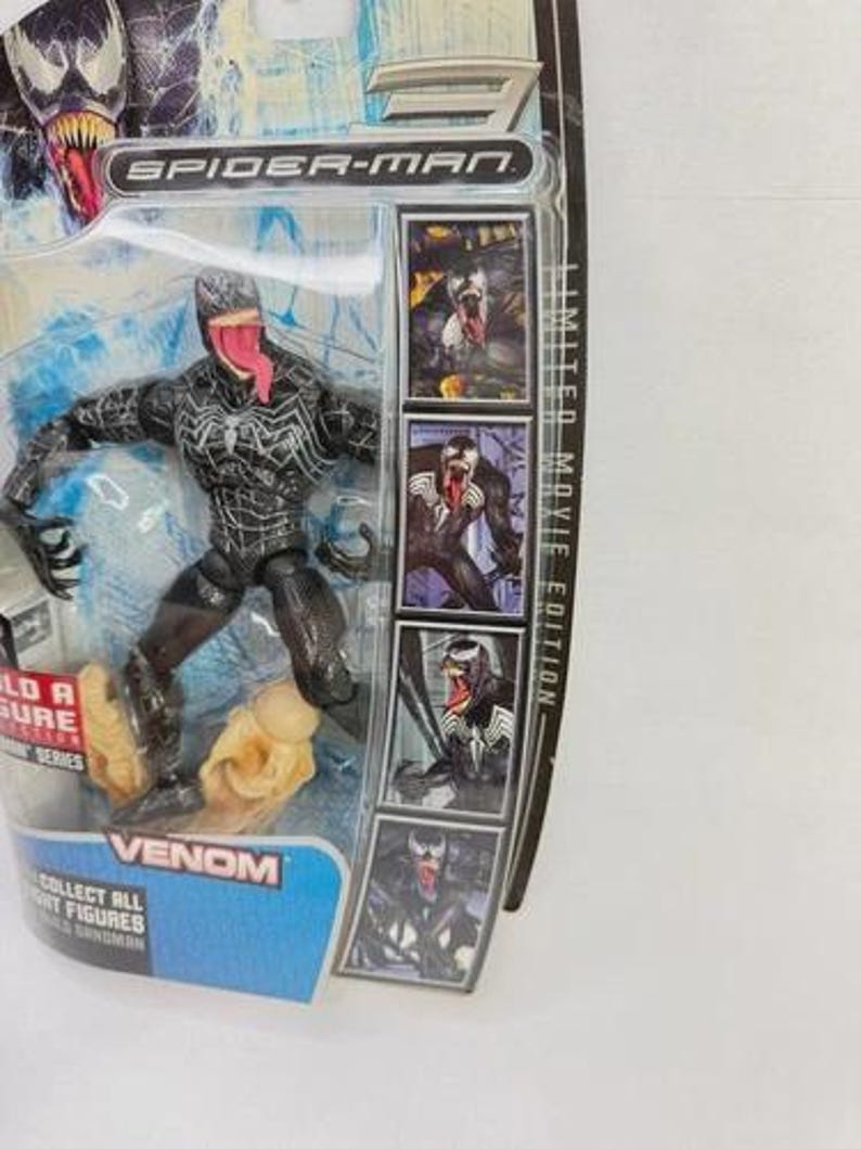 May include: A Venom action figure from the Spider-Man movie series, in its original packaging. The figure is black with white accents and a long red tongue. The packaging includes the text "VENOM" and "COLLECT ALL FIGHT FIGURES".