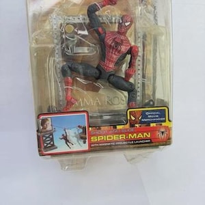 May include: Spider-Man action figure in a clear plastic package. The figure is red and black, posed in a dynamic action stance. The packaging includes the text "SHOOT AND SLIDE SPIDER-MAN" and "OFFICIAL MOVIE MERCHANDISE".