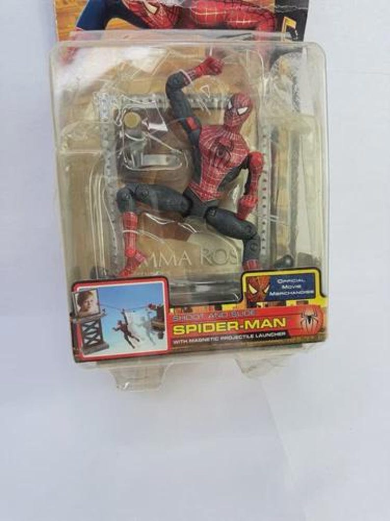 May include: Spider-Man action figure in a clear plastic package. The figure is red and black, posed in a dynamic action stance. The packaging includes the text "SHOOT AND SLIDE SPIDER-MAN" and "OFFICIAL MOVIE MERCHANDISE".