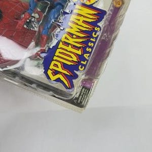 May include: A Spider-Man action figure in a clear plastic package. The figure is red and blue, with the words "Spider-Man Classics" in yellow and blue. The package also includes a purple accessory.
