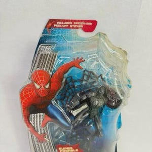 Peut inclure: Figurines de Spider-Man dans un emballage plastique. Une figurine est rouge et bleue, l'autre est noire. L'emballage comprend le texte "INCLUDES SPIDER-MAN PEEL-OFF STICKER" et "SUPER POSABLE WEB!"