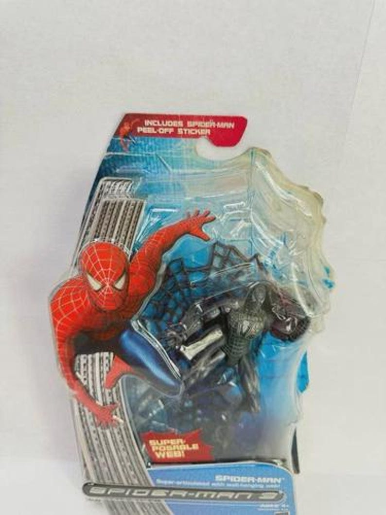 Peut inclure: Figurines de Spider-Man dans un emballage plastique. Une figurine est rouge et bleue, l'autre est noire. L'emballage comprend le texte "INCLUDES SPIDER-MAN PEEL-OFF STICKER" et "SUPER POSABLE WEB!"