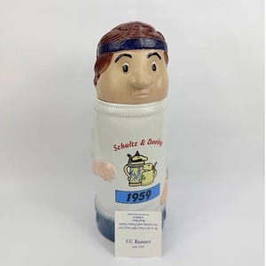 Pode incluir: Uma caneca de cerâmica em forma de corredor com cabelo castanho e uma faixa azul. O corpo branco apresenta o texto Schultz & Dooley, uma ilustração de duas canecas de cerveja e o ano 1959 em azul. Um pequeno cartão branco está anexado.