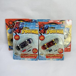 Puede incluir: Varios fingerboards empaquetados con gráficos de Spider-Man. El embalaje muestra "The Amazing Spider-Man" y "Finger Board Pro Model Skateboards." Cada paquete incluye un monopatín en miniatura y herramientas.