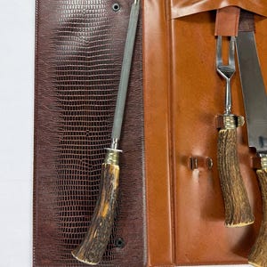 May include: A brown leather case with a textured, reptile-like pattern, open to reveal a sharpening steel, a fork, and two knives. The handles of the utensils are made of a light brown, antler-like material.