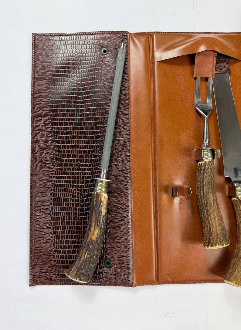 May include: A brown leather case with a textured, reptile-like pattern, open to reveal a sharpening steel, a fork, and two knives. The handles of the utensils are made of a light brown, antler-like material.