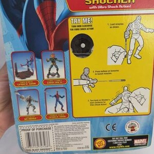 May include: A blue and red Spider-Man action figure package. The packaging features the words "DUAL BLAST SHOCKER" and "TRY ME!". The package also includes illustrations of Spider-Man and other characters.