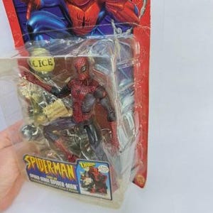 Peut inclure: Figurine de Spider-Man dans un emballage plastique. La figurine est rouge et noire, avec un graphisme Spider-Man bleu et rouge sur l'emballage. L'emballage porte &eacute;galement les mots "Spider-Man" et "THWIP!"