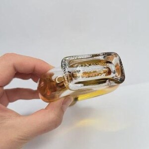 May include: A clear glass bottle with a rectangular base and rounded body, filled with a golden liquid. The bottle is held at an angle, showcasing its shape and contents against a white background.