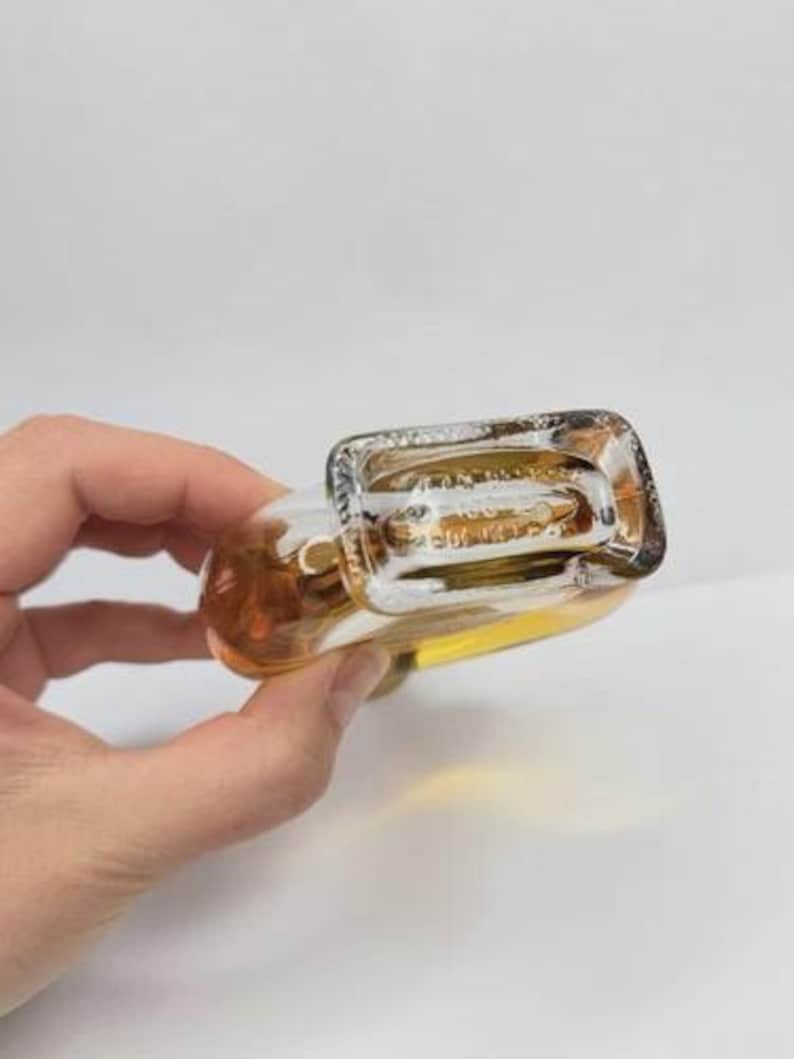 May include: A clear glass bottle with a rectangular base and rounded body, filled with a golden liquid. The bottle is held at an angle, showcasing its shape and contents against a white background.