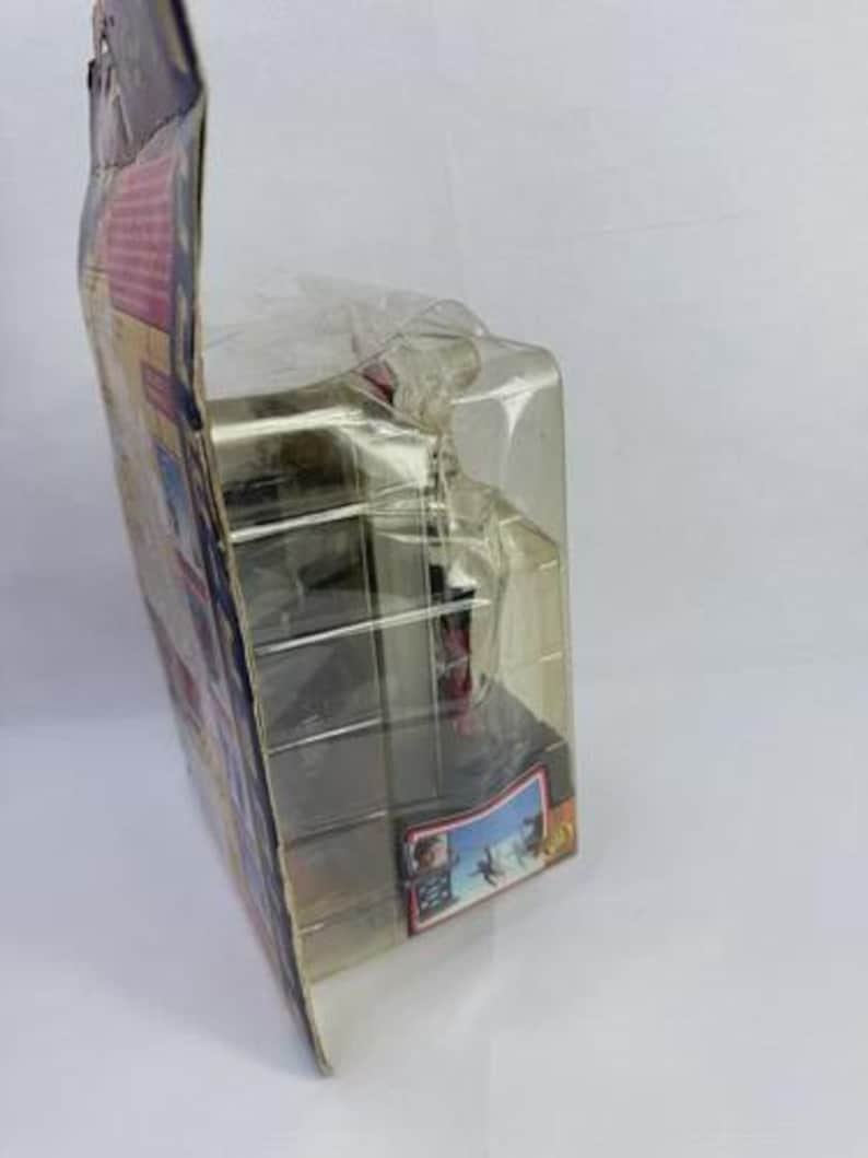 May include: A packaged action figure in a clear plastic case. The packaging has colorful graphics and text. The figure is partially visible inside the case. The case is sealed and appears to be new.