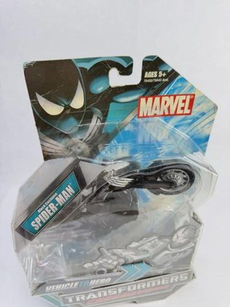 Puede incluir: Un juguete Marvel Spider-Man Black Costume Vehicle to Hero Transformers. El juguete es negro y plateado, empaquetado en una funda de pl&aacute;stico transparente con un respaldo de cart&oacute;n azul y negro de la marca Marvel. El embalaje indica que el juguete es para mayores de 5 a&ntilde;os.