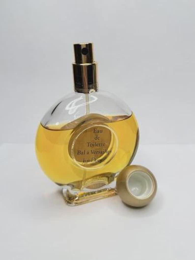 May include: A clear, round perfume bottle with a gold-colored sprayer and a matching cap. The bottle contains a golden-yellow liquid and has a label that reads "Eau de Toilette Bal &agrave; Versailles Jean Desprez."