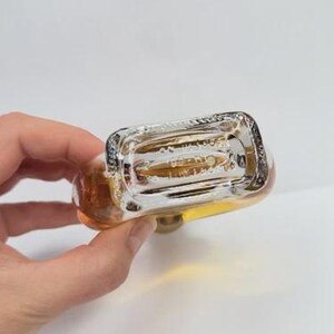 May include: A clear glass bottle with a rectangular base and rounded top, filled with a golden liquid. The bottle has a silver-colored cap and is held against a white background. The bottom of the bottle is visible.