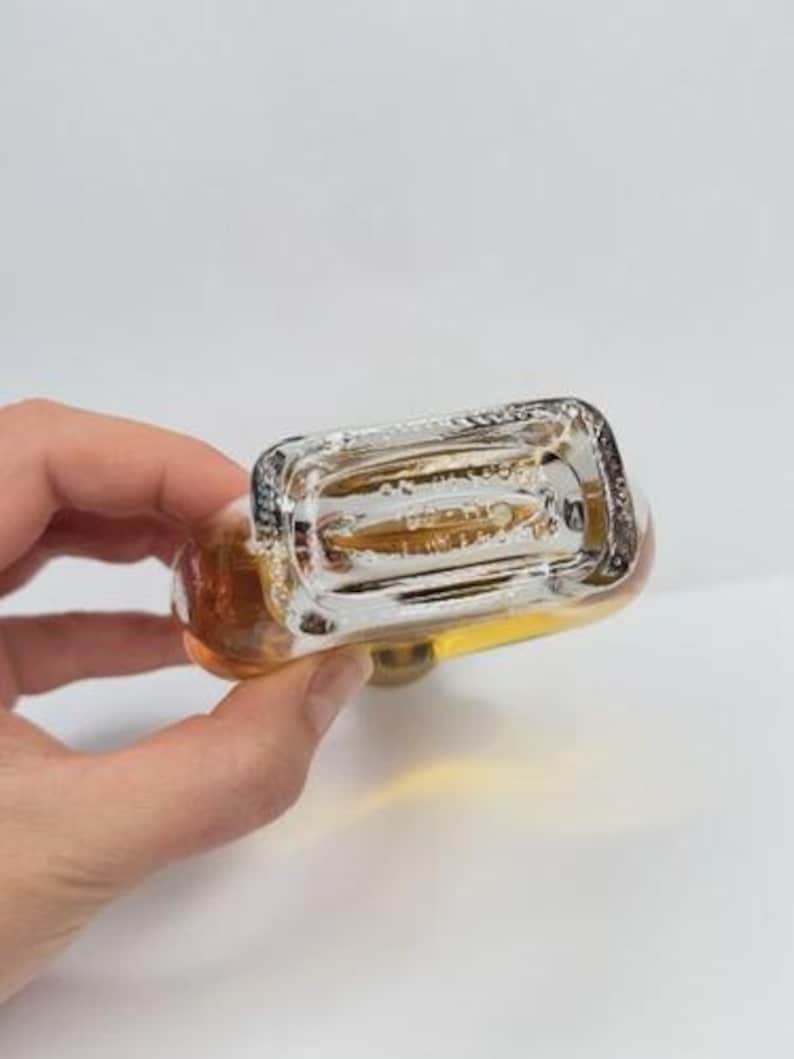 May include: A clear glass bottle with a rectangular base and rounded top, filled with a golden liquid. The bottle has a silver-colored cap and is held against a white background. The bottom of the bottle is visible.