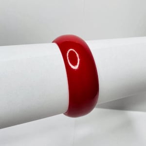 May include: A glossy, solid red bangle bracelet. The bracelet is a thick, rounded band, and the color is a vibrant, saturated red. The bracelet is displayed on a white background. This accessory is suitable for everyday wear.