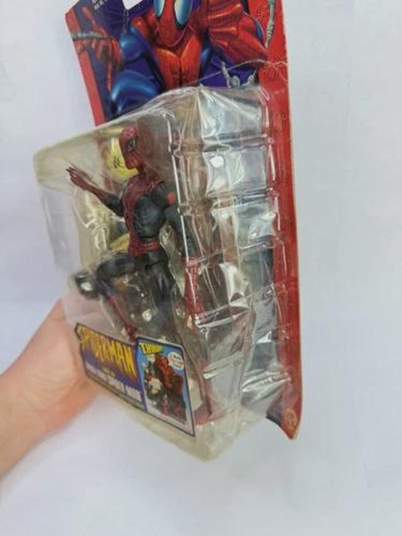Peut inclure: Figurine de Spider-Man dans un emballage plastique. La figurine est rouge et noire, avec un costume d&eacute;taill&eacute;. L'emballage a un fond rouge et bleu avec le logo Spider-Man et des illustrations du personnage. La figurine mesure environ 15 cm de haut.