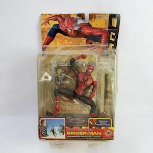 May include: A Spider-Man action figure in a clear plastic package. The figure is red and black, in a dynamic pose. The packaging includes the text "SHOOT AND SLIDE SPIDER-MAN" and "OFFICIAL MOVIE MERCHANDISE".