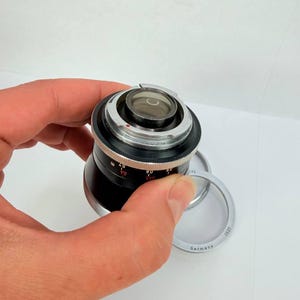 Puede incluir: Un objetivo de c&aacute;mara negro y plateado con un elemento de vidrio transparente. El objetivo tiene marcas de medici&oacute;n y est&aacute; sostenido por una mano. Tambi&eacute;n es visible un anillo plateado con el texto "Germany".