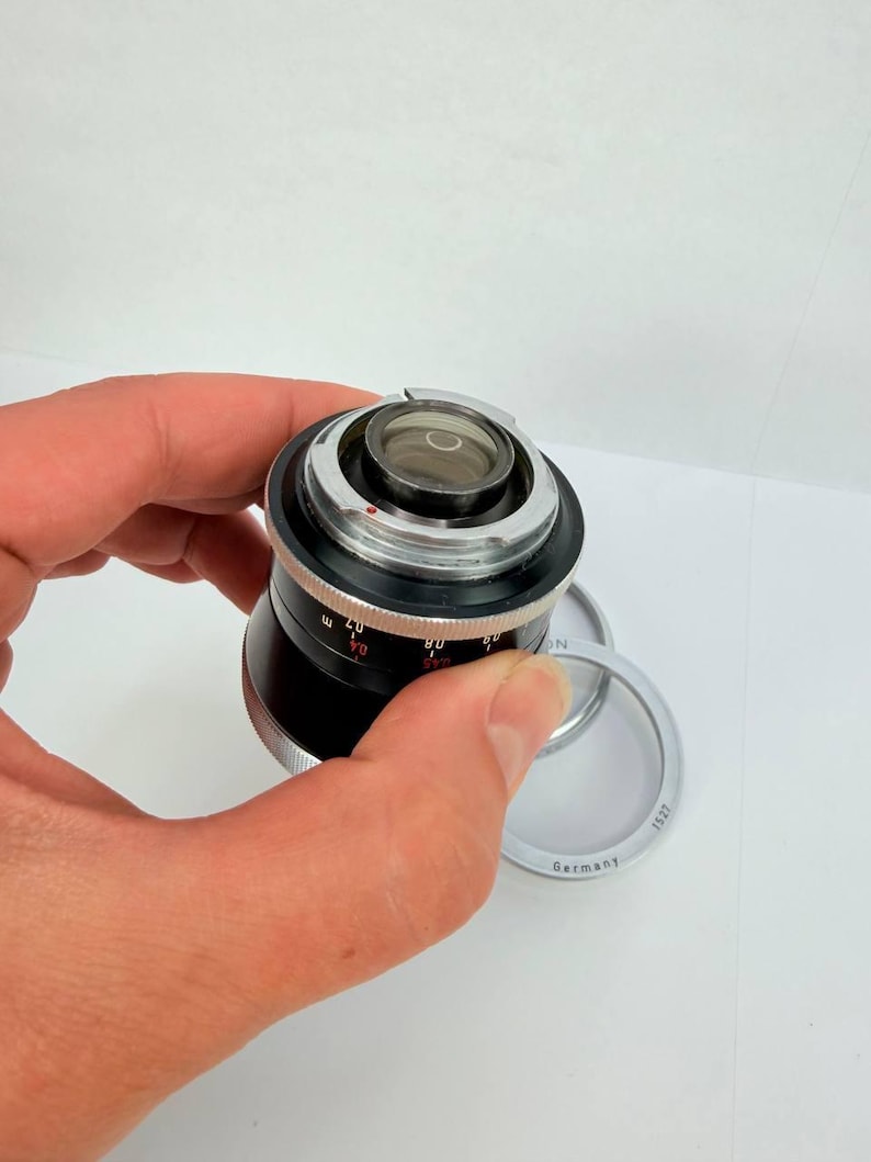 Puede incluir: Un objetivo de c&aacute;mara negro y plateado con un elemento de vidrio transparente. El objetivo tiene marcas de medici&oacute;n y est&aacute; sostenido por una mano. Tambi&eacute;n es visible un anillo plateado con el texto "Germany".