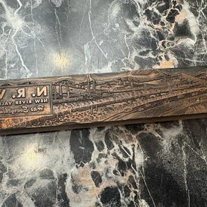 May include: A rectangular wooden printing block featuring a relief engraving of an industrial landscape with smokestacks and railway tracks. The text N. R. V. NEW RIVER VALLEY Has Everything is embossed on the left side in a clear, sans-serif font.