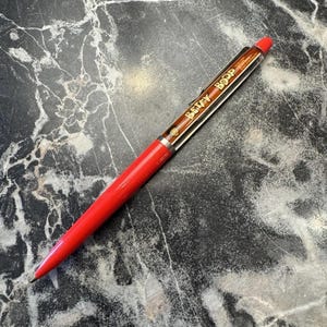 Könnte beinhalten: Ein roter Stift mit dem Text "Betty Boop" in goldfarbenen Buchstaben. Der Stift hat einen roten Schaft und eine silberne Spitze. Er liegt auf einer schwarz-grauen Marmoroberfläche.