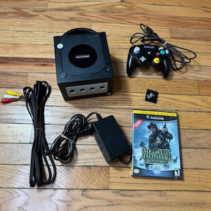 Può includere: Una console Nintendo GameCube nera con controller nero abbinato, un alimentatore, un cavo AV, una scheda di memoria con le lettere P.M. scritte sopra e il videogioco Medal of Honor Frontline nella sua custodia originale.